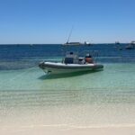 A small rubber duck anchored in shallow blue water with small boats in the background